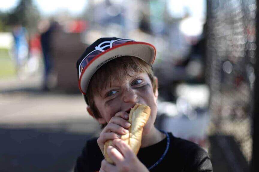 Little boy in a baseball cap eating a hot dog at a game