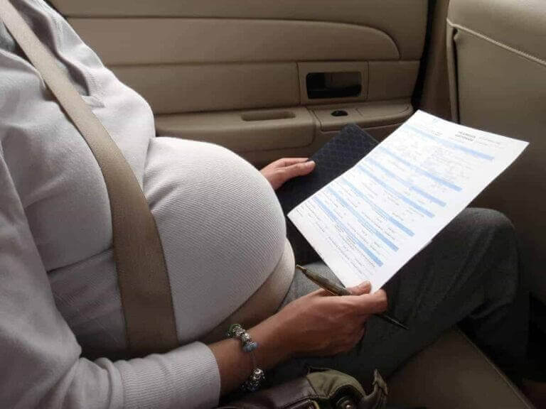 A pregnant woman wearing a seatbelt in a taxi looking at paperwork from the doctor.