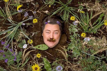 Comedian mimics growing out of the dirt with only his face showing surrounded by wildflowers