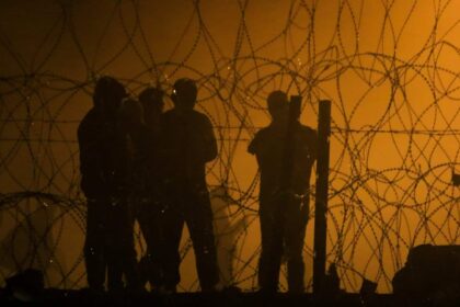 Migrants looking through a barbed wire fence as the sun rises in an orange sky