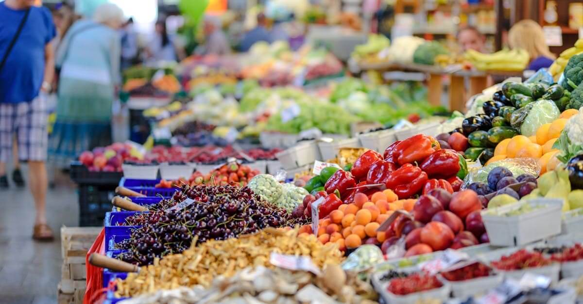 Tariffs affect food prices. Colorful assortment of fresh fruits and vegetables in a bustling outdoor market scene.