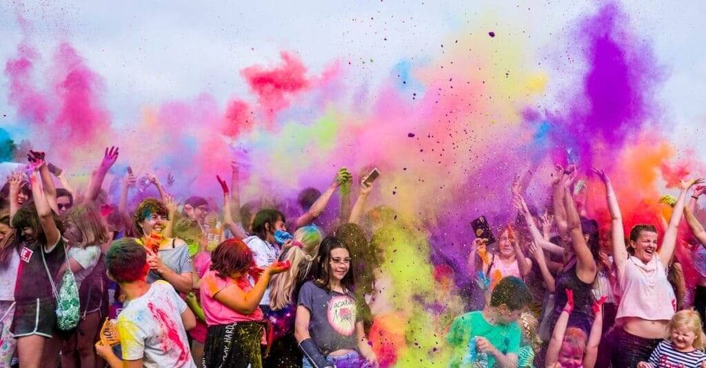 A joyful crowd covered in vivid colors celebrates a festival outdoors with flying powder.