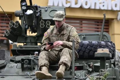 A soldier in camouflage resting on an armored vehicle, using a smartphone outside a McDonald's.